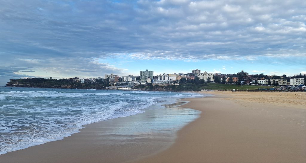 A quiet morning view of a sandy beach with gentle waves rolling in under a cloudy blue sky. In the distance, a row of coastal buildings lines the shore, with green hills behind them.