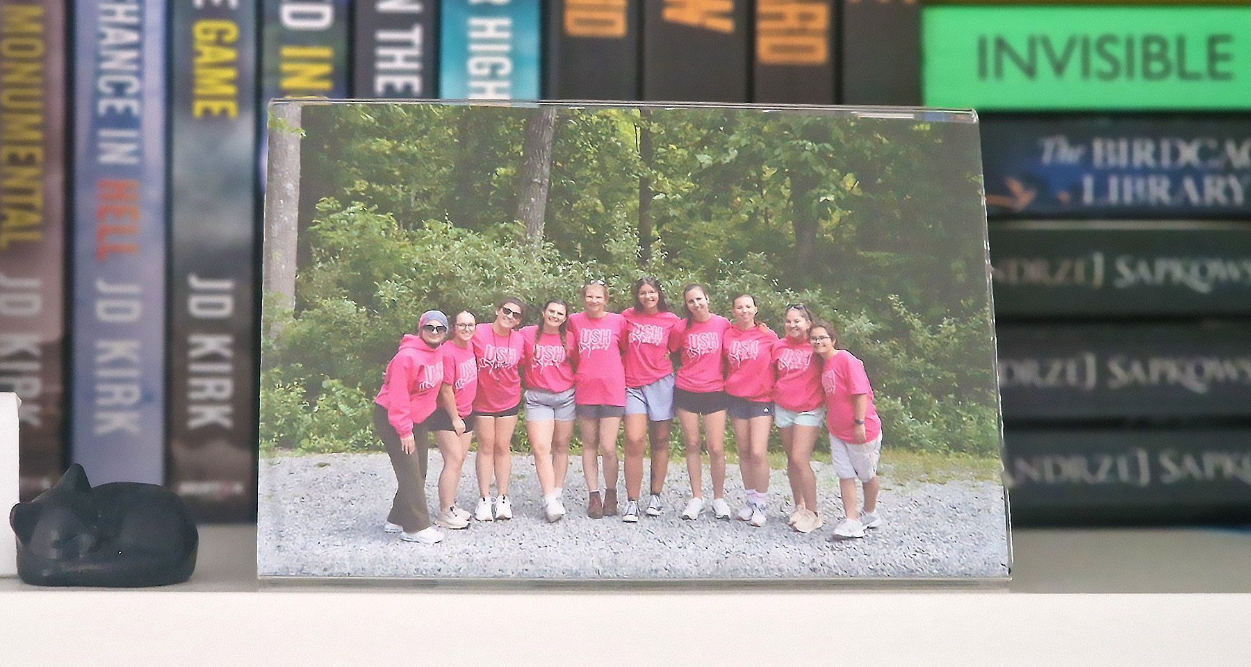 A photo frame displays a group of ten young women in bright pink USHthis shirts and hoodies standing outdoors with their arms around each other. The frame is placed on a bookshelf with the books out of focus in the background.